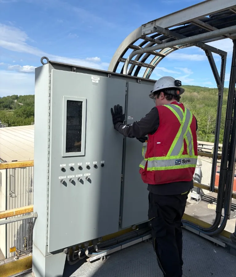 Electrician in hard hat and safety vest inspecting an outdoor industrial electrical panel